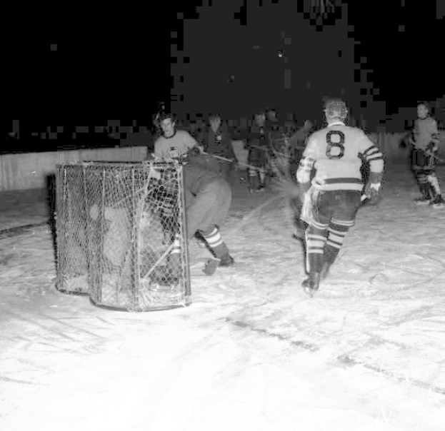 Schwarzes und weißes Foto von Männern, die Hockey auf einem Eisplatz mit einem Netz im Vordergrund und einer Wand im Hintergrund spielen