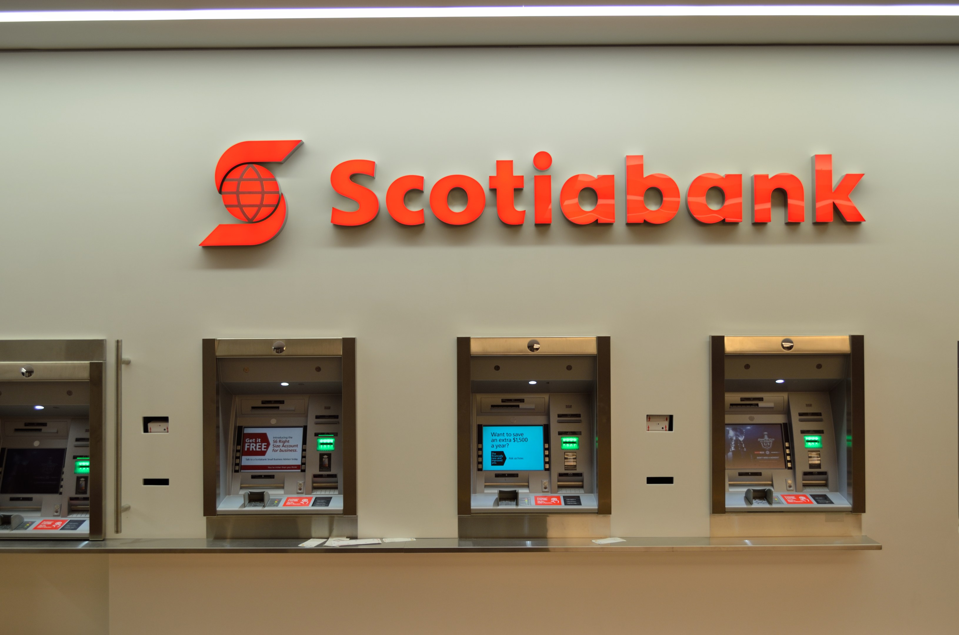 Ein modernes Scotiabank-Geldautomatengerät mit schwarzer und silberner Farbgebung, das eine große Anzeigebildschirm und Steuerungsknöpfe aufweist und vor einer hellgrauen Wand mit gerahmten Bildern und dem Text "Scottish Bank" im Hintergrund positioniert ist.