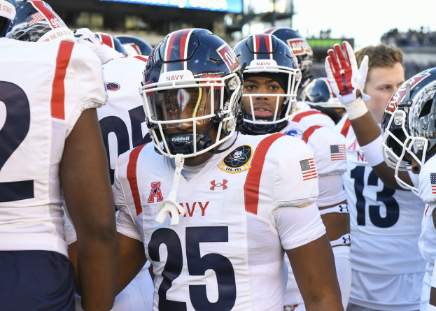 Gruppe von Männern in Fußballausrüstung auf einem Feld mit Helmen und Handschuhen, mit einer unscharfen Menge, einem Anzeigebrett mit der Aufschrift "Auburn vs Virginia Tech - 11/28/15 College Football Pick, Odds, and Prediction" und Himmel im Hintergrund.