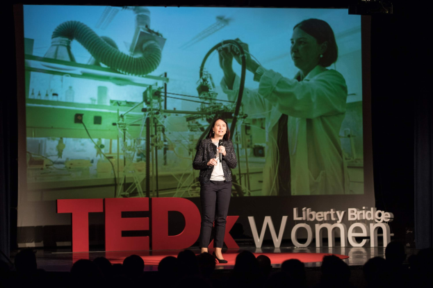 Frau auf einer Bühne mit Mikrofon, hinter ihr ein Display mit Bild einer Frau und Text zum TEDxWomen-Event.