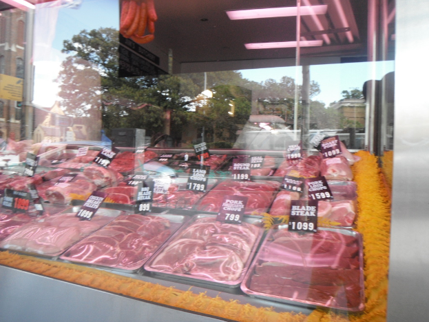 Eine Glasvitrine in einem Fleischgeschäft, die Tabletts mit Fleisch und Preisschildern zeigt, mit Gebäuden und Bäumen im Hintergrund und der Decke des Raums oben im Bild.