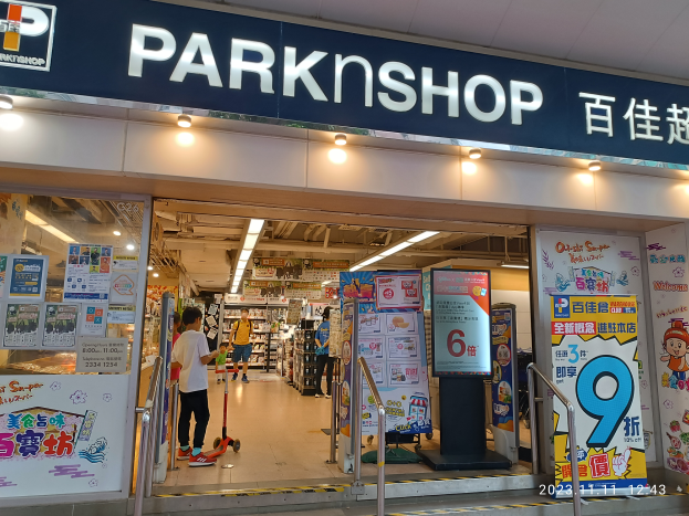 Ein Parkshop in einem Einkaufszentrum mit einem Schild, auf dem 'Park Shop' steht, und einer Gruppe von Menschen auf dem Boden, umgeben von Metallpfosten, Schildern, Bannern, Gestellen mit Gegenständen und Deckenleuchten.