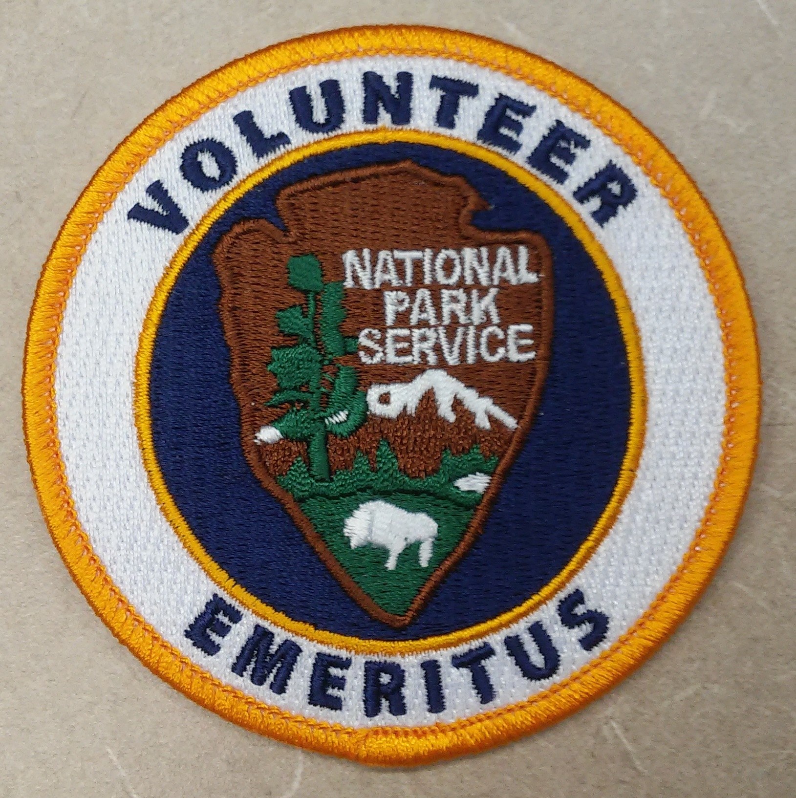 Runder Flicken mit blauem Rand und weißem Hintergrund, der mittig den schwarzen Text "Volunteer National Park Service Emeritus" in einem gelben Kreis zeigt.