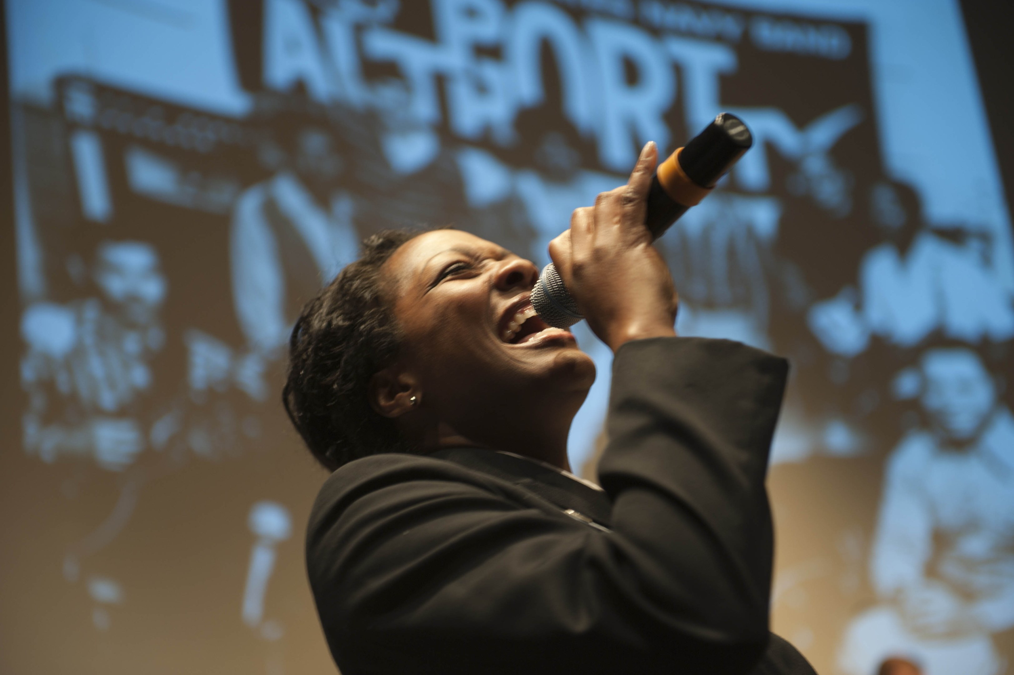 Eine Frau in einem schwarzen Kleid mit einem Dutt singt leidenschaftlich in ein Mikrofon auf der Bühne, ihr Gesicht von Scheinwerfern beleuchtet und ihre Augen vor Emotion geschlossen.