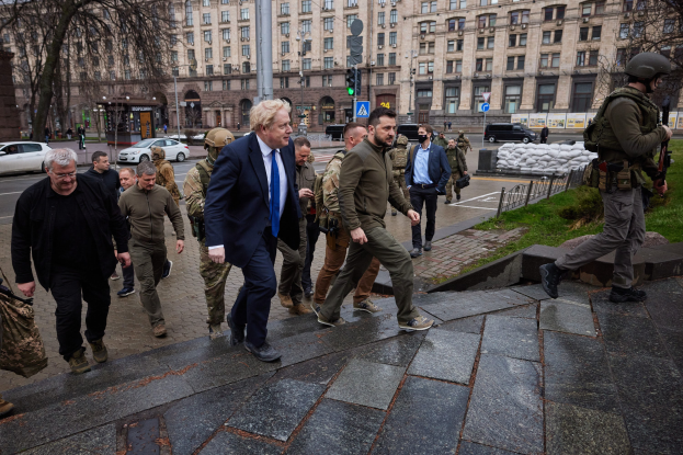 Eine Gruppe von Menschen, darunter Boris Johnson, geht auf einem Bürgersteig neben einem Gebäude, einige tragen Helme und halten Waffen, mit Fahrzeugen, Laternenmasten, Verkehrsampeln, Bäumen und Gebäuden im Hintergrund.