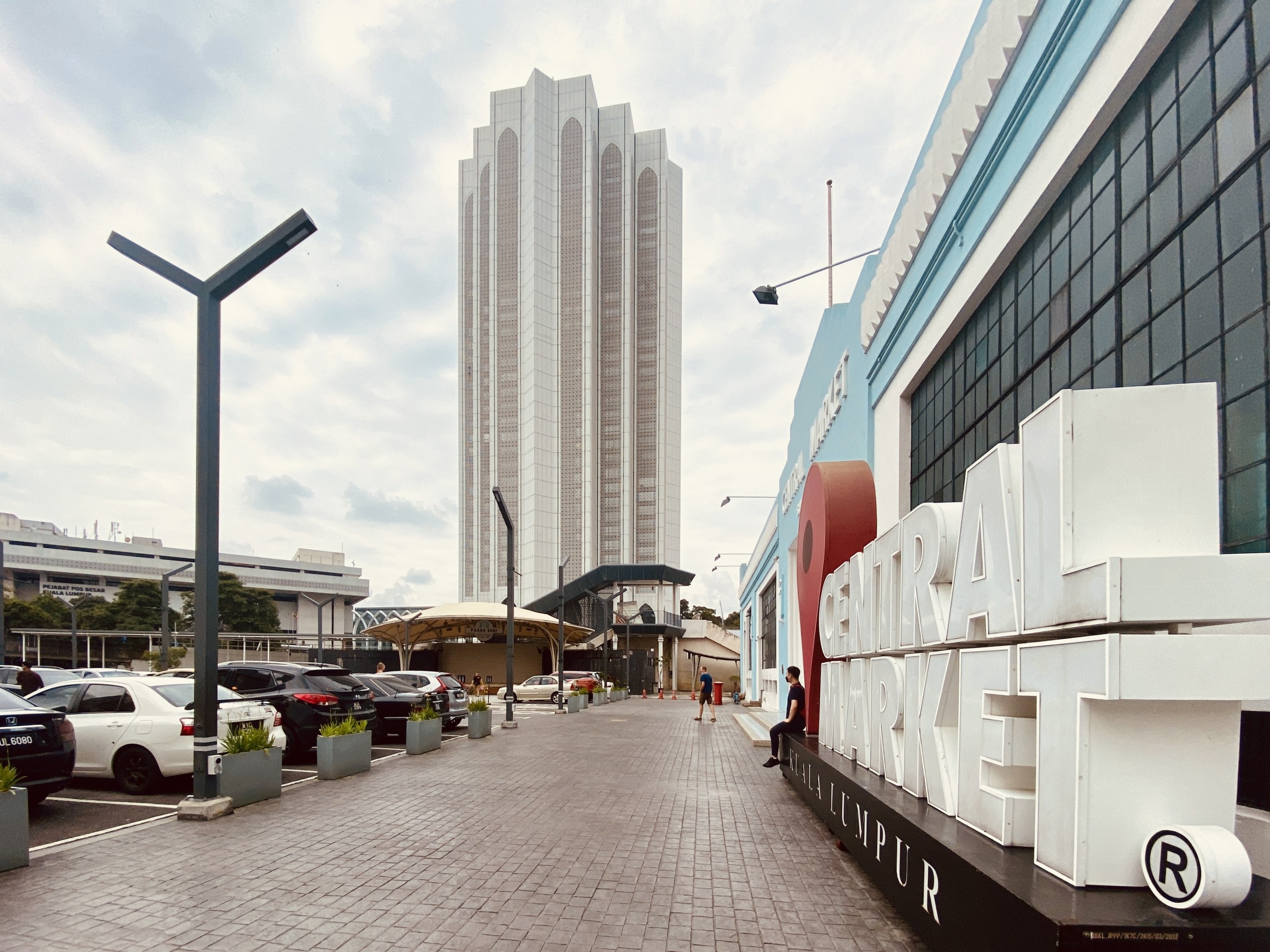 Ein belebter zentraler Markt in Kuala Lumpur, Malaysia, mit hohen Gebäuden, Straßenlaternen, Straßenlaternen, Kraftfahrzeugen, Fußgängern auf dem Gehweg, Namensschildern, Bäumen und einem bewölkten Himmel.