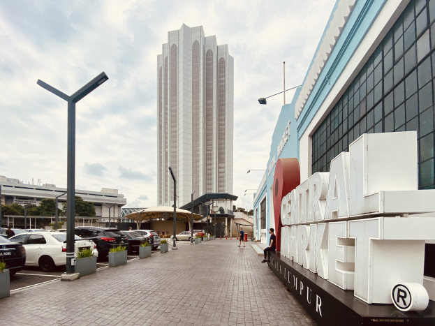 Ein belebter zentraler Markt in Kuala Lumpur, Malaysia, mit hohen Gebäuden, Straßenlaternen, Straßenlaternen, Kraftfahrzeugen, Fußgängern auf dem Gehweg, Namensschildern, Bäumen und einem bewölkten Himmel.