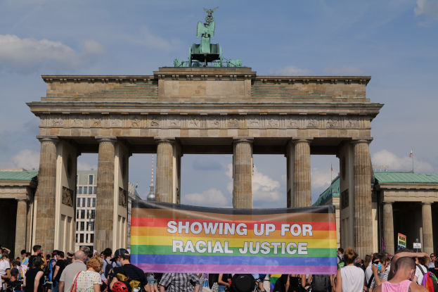 Eine Gruppe von Menschen steht vor dem Brandenburger Tor in Berlin, Deutschland, mit einer Tafel, auf der 'Rassengerechtigkeit' steht, im Hintergrund sind die Säulen des Tors, eine Statue, Gebäude und ein bewölkter Himmel zu sehen.