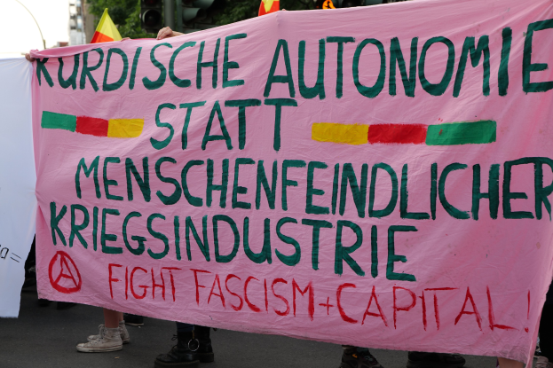 Eine Gruppe von Menschen hält eine pinkfarbene Fahne mit der Aufschrift 'Kurdische Autonomie Statt Menschenfeindlicher Kriegsindustrie Fight Fascism Capital' auf einer Straße mit Verkehrszeichen, Fahnen, Bäumen, Gebäuden und einem klaren blauen Himmel im Hintergrund.