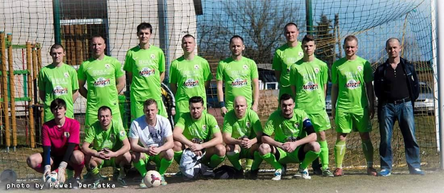 Gruppe von Männern in grünen T-Shirts auf einem Fußballfeld mit einem Ball und einem Torpfosten, mit Bäumen, Gebäuden und Fahrzeugen im Hintergrund.