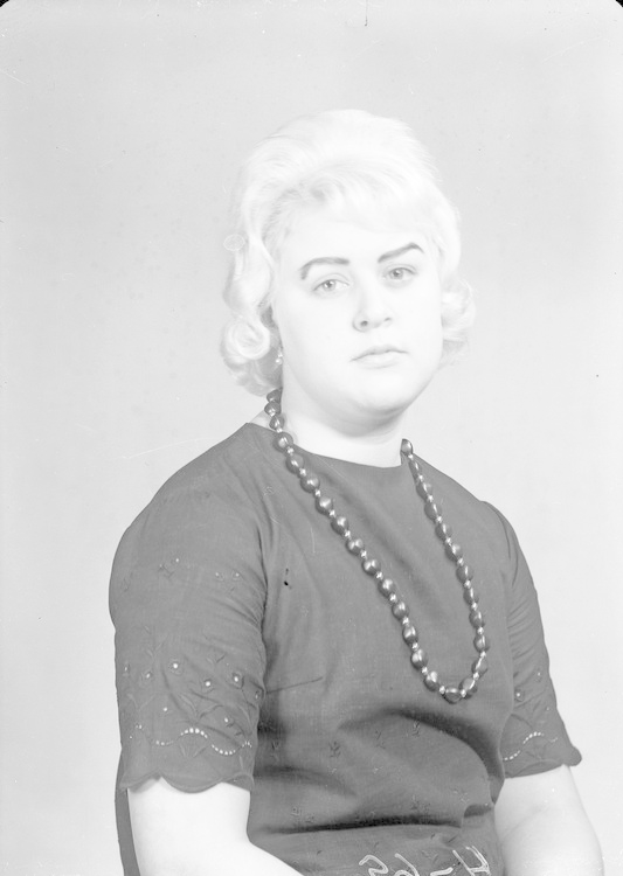 Ein Schwarz-Weiß-Foto einer Frau mit ernstem Gesichtsausdruck, deren Haar zurückgebunden ist und die eine Kette und ein einfaches Kleid mit hohem Kragen und langen Ärmeln trägt.