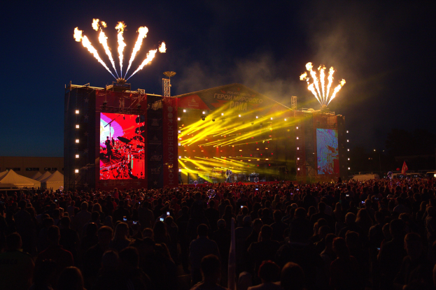Eine große Menschenmenge bei einem Musikfestival schaut auf eine hell erleuchtete Bühne, mit Zelten links und Feuerwerk am dunklen Himmel dahinter.