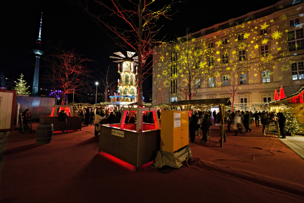 Ein geschäftiges Weihnachtsmarkt-Szenario in Berlin, Deutschland, mit Menschen um dekorierte Stände, festliche Lichter, Tannenbäume, Gebäude, Laternenmäste und einen Turm unter einem dunklen Himmel.