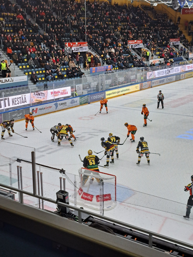 Eishockeyspieler konkurrieren auf einem Eisring mit einem Netz und einer umliegenden Umzäunung, während Zuschauer von Stühlen aus zusehen und Banner im Hintergrund unter Stadionbeleuchtung hängen.