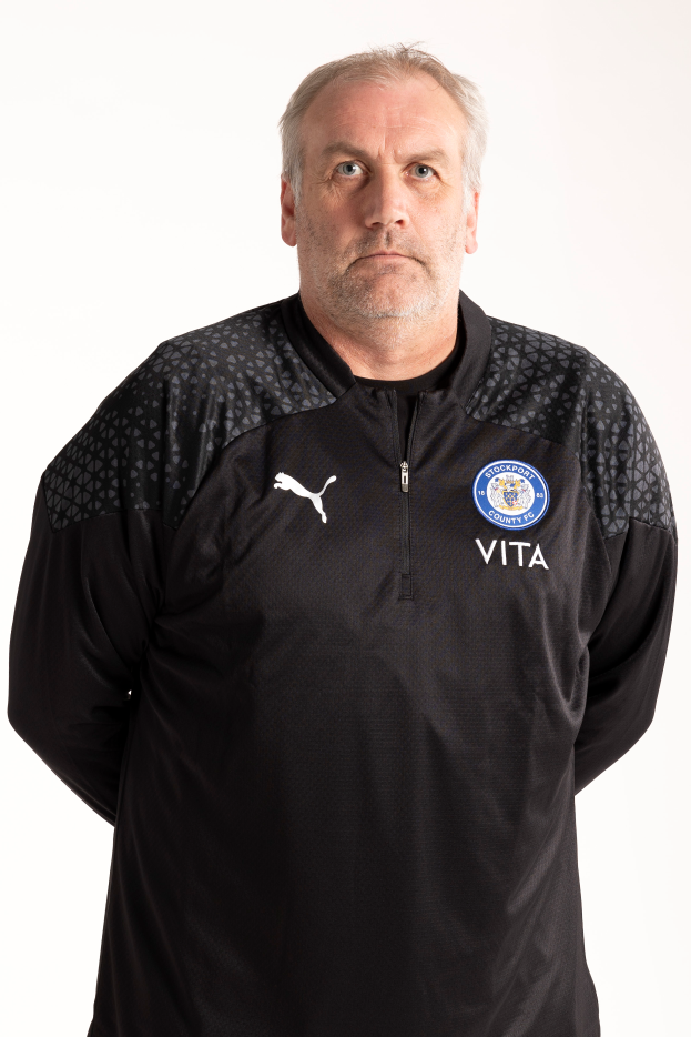 Ein Mann in einer schwarzen Jacke steht vor einem weißen Hintergrund und posiert mit ernster Miene, wobei er wie ein Fussballtrainer aussieht.