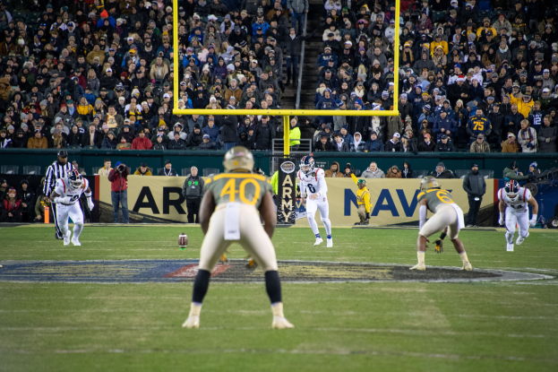 Fußballspieler in Helmen und Uniformen auf einem Feld mit Zuschauern auf den Stadionrängen, umgeben von Torpfosten und Beschilderung.