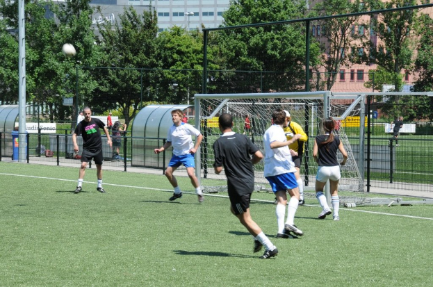 Menschen in Kleidung beim Fußballspielen auf einem Feld mit einem Zaun und einem Tor in der Mitte, einem Pfosten und einem Fußball oben links und Bäumen und Gebäuden im Hintergrund.