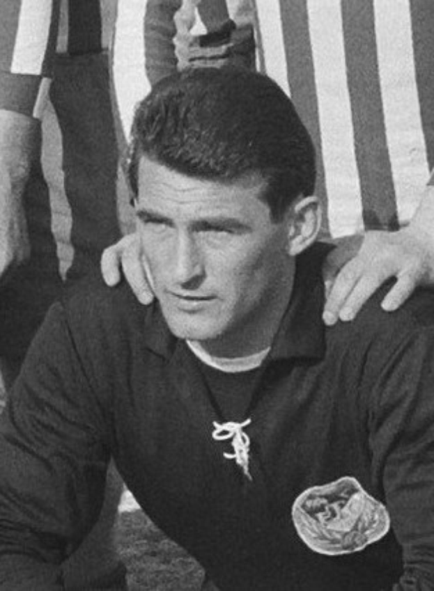 Ein Schwarz-Weiß-Foto eines Mannes, der auf einem grünen Fussballfeld sitzt und ein T-Shirt sowie eine Kette trägt, mit zwei Personen dahinter.