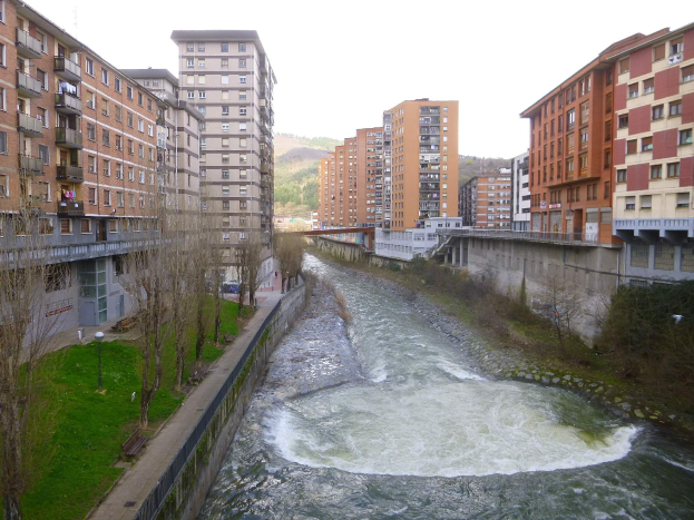 Ein Fluss fließt durch eine städtische Gegend mit hohen Gebäuden, umgeben von grünem Gras, Bäumen und Laternenmasten, mit einer Brücke darüber; Hügel und ein klarer blauer Himmel sind im Hintergrund zu sehen.