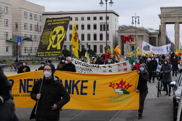 Eine große Gruppe von Menschen marschiert mit Schildern und Fahnen auf einer Straße während einer Demonstration gegen Atomkraft in Deutschland, mit Fahrzeugen auf der rechten Seite und Gebäuden, Laternen, Bäumen und einem Bogen im Hintergrund.