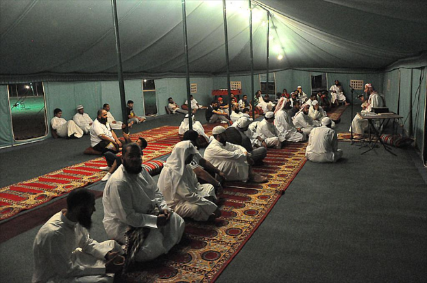 Eine Gruppe von Menschen sitzt auf dem Boden in einem Camp, mit einer Person auf einem Stuhl rechts, einem Mikrofon davor, einem Tisch daneben und einer Lampe oben im Bild.