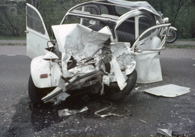 Ein verunglücktes Auto mit schwerer Frontschaden auf der Seite einer Straße, mit Bäumen, einem Gebäude und einem anderen Fahrzeug im Hintergrund.
