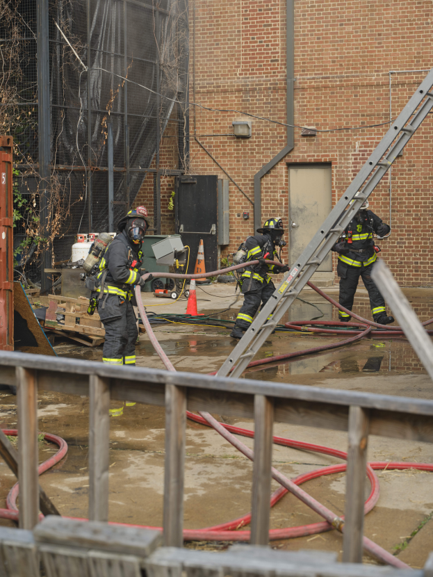 Eine Gruppe von Feuerwehrleuten in Helmen, die Rohre halten, arbeiten daran, ein Gebäude Feuer zu löschen, mit verschiedenen Ausrüstungen und Strukturen drumherum, einschließlich eines Metallzauns, Rohren auf dem Boden, einem Container, einem Verkehrskegel, einem Baum und dem Himmel im Hintergrund.