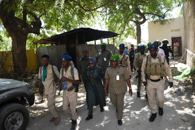 Gruppe von Polizisten in Uniform, die eine Straße entlanggehen, einige halten Waffen, mit Bäumen, Schuppen und Gebäuden im Hintergrund.