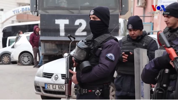 Eine Gruppe von Polizisten in Uniform steht mit Waffen auf einer Straße während einer Anti-Regierungsdemo in Ankara, Türkei, mit Fahrzeugen, Menschen, Gebäuden und Texttafeln im Hintergrund.