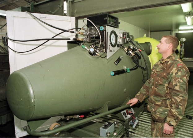 Ein Mann in militärischer Uniform steht neben einer großen grünen Bombe in einer unordentlichen industriellen Umgebung mit Maschinen, Drähten und beleuchtetem Hintergrund.