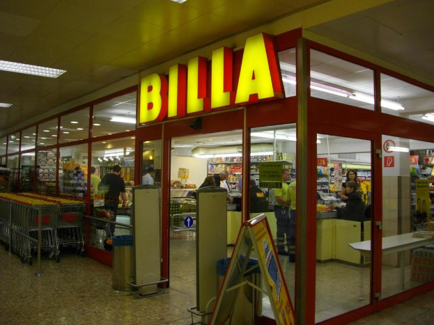 Ein Supermarkt mit einem Eingang in der Mitte, Mülltonnen daneben, eine Reihe von Einkaufswagen auf der linken Seite, Deckenbeleuchtung und einige Menschen, die Produkte anschauen.