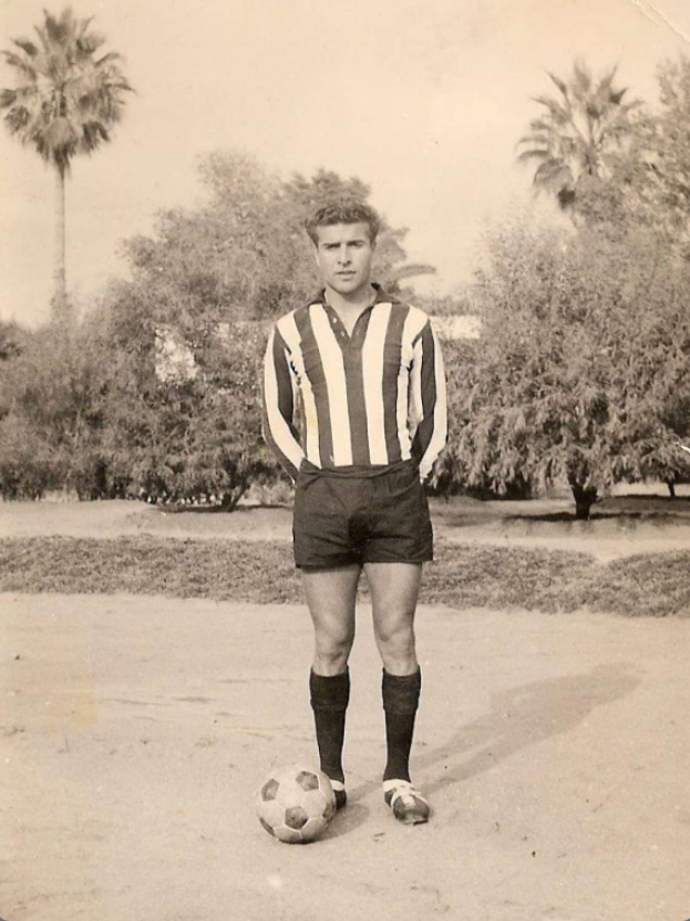 Ein Schwarz-Weiß-Foto eines älteren Mannes mit einem Fußball, der draußen vor einem Baum und dem Himmel steht.