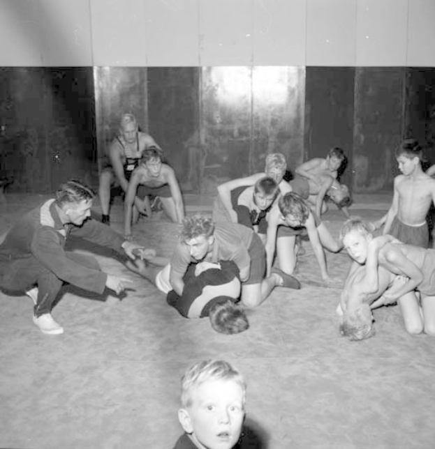 Schwarz-weißes Bild von jungen Jungs, die in einem Gymnastikraum miteinander ringen, mit einer Wand im Hintergrund.