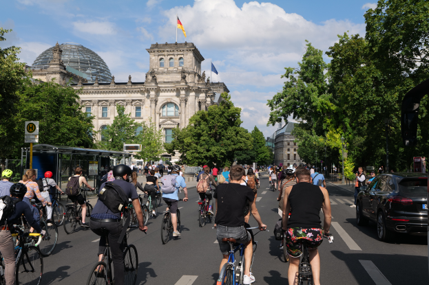 Eine Gruppe von Menschen, die auf Fahrrädern eine von Bäumen gesäumte Straße in Berlin, Deutschland, entlangfahren, mit Gebäuden auf beiden Seiten, einer Bushaltestelle auf der rechten Seite und einer Flagge, die auf einem der Gebäude weht, unter einem bewölktem Himmel.