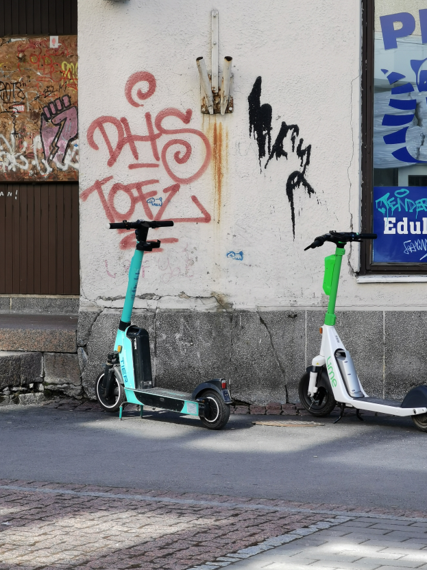 Zwei Elektro-Scooter nebeneinander auf einer Straße geparkt, mit einer Graffiti-bedeckten Wand und einem Plakat an einem Fenster im Hintergrund.