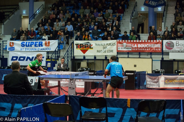 Indoor-Tischtennis-Match mit zwei Spielern und einigen stehenden Zuschauern, sitzendem Publikum, leeren Stühlen und Werbetafeln in einer Stadionatmosphäre.