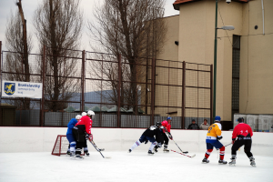 Menschen beim Eishockeyspielen auf einer Eisfläche mit Gebäuden, Bäumen, einer Straßenlaterne, einem Namensschild und Zäunen im Hintergrund unter einem Himmel.