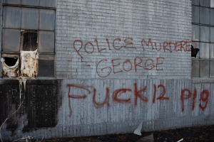 Graffiti an einer Hauswand mit der Aufschrift "Polizei ermordet George Fuck 12 P19" mit Fenstern und verstreuten Gegenständen davor.