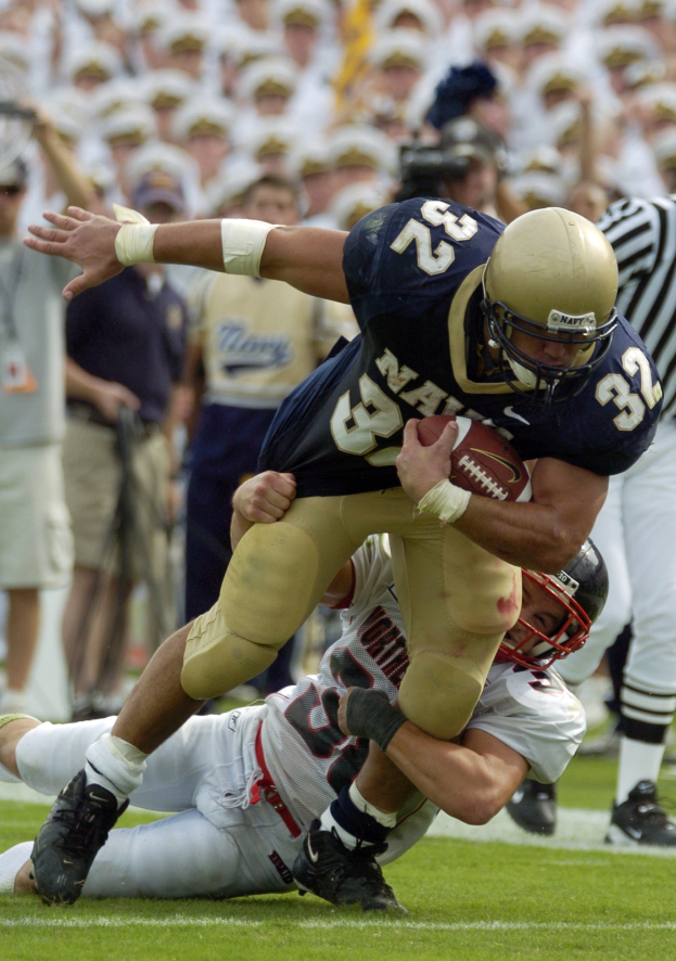 Zwei Fußballspieler im Einsatz, einer in Sportkleidung und Helm, der von einem anderen auf dem Feld getackelt wird, mit anderen und Zuschauern im Hintergrund.
