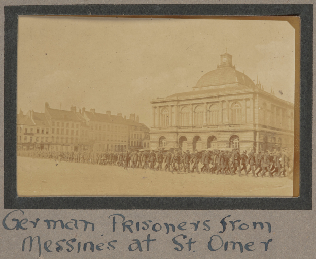 Deutsche Kriegsgefangene aus Messines vor einem Gebäude mit Text unten gegen einen klaren blauen Himmel.