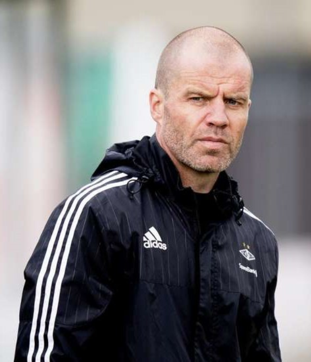 Mann mit Glatze in marineblauer Jacke mit weißen Streifen und weißem Kragen, der auf einem Fußballfeld steht und die Hände gefaltet hat.