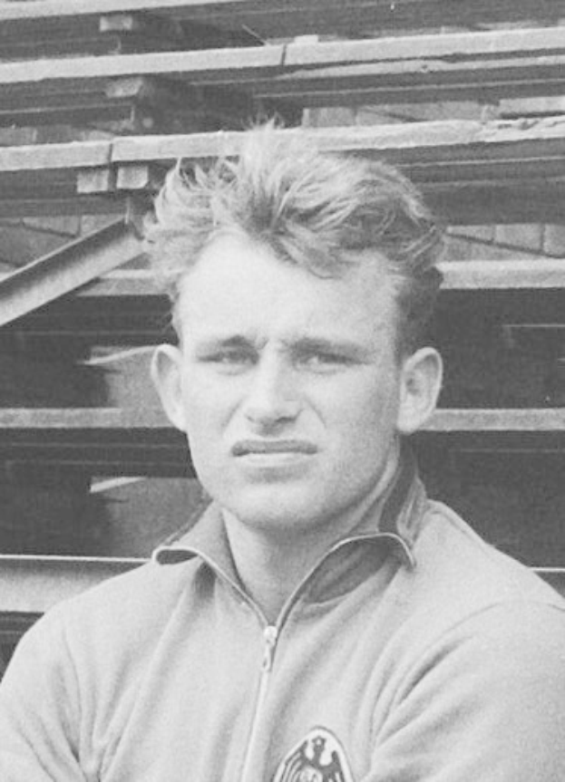 Ein Schwarz-Weiß-Foto eines älteren Mannes in einer Jacke vor einem Haufen Holzplanken.