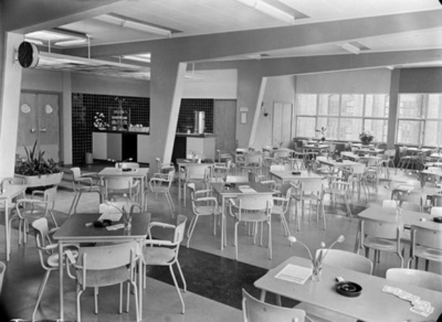 Schwarzes und weißes Foto einer Cafeteria mit Tischen und Stühlen, Tellern, Besteck, Gläsern, Vasen und Deckenleuchten sowie Türen und Fenstern im Hintergrund.