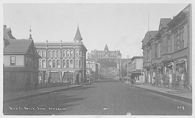 Ein Schwarz-Weiß-Foto einer Stadtstraße mit Gebäuden auf beiden Seiten, Masten, die die Straße säumen, und einem klaren Himmel im Hintergrund, mit Text unten, der "Third St North von University" lautet.