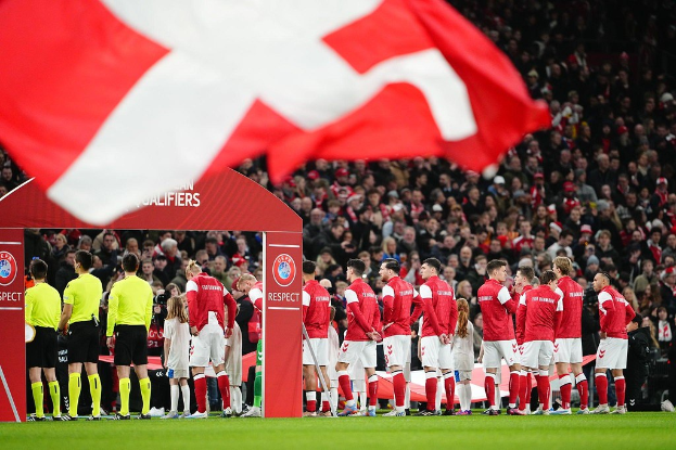 Eine Gruppe von Menschen auf einem Fußballfeld mit einer roten und weißen Fahne im Vordergrund, einem Bogen mit Text "Bayern München vs. Bayern München Wetten & Vorschau" im Hintergrund und einer großen Menschenmenge im Stadion.