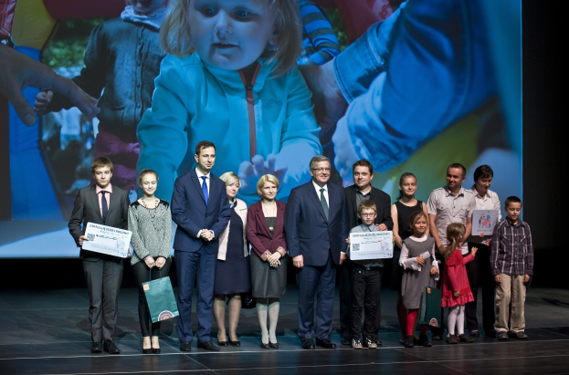 Gruppe von Menschen auf einer Bühne mit Taschen und Urkunden, im Hintergrund ein Bildschirm mit einem Kind und anderen Personen