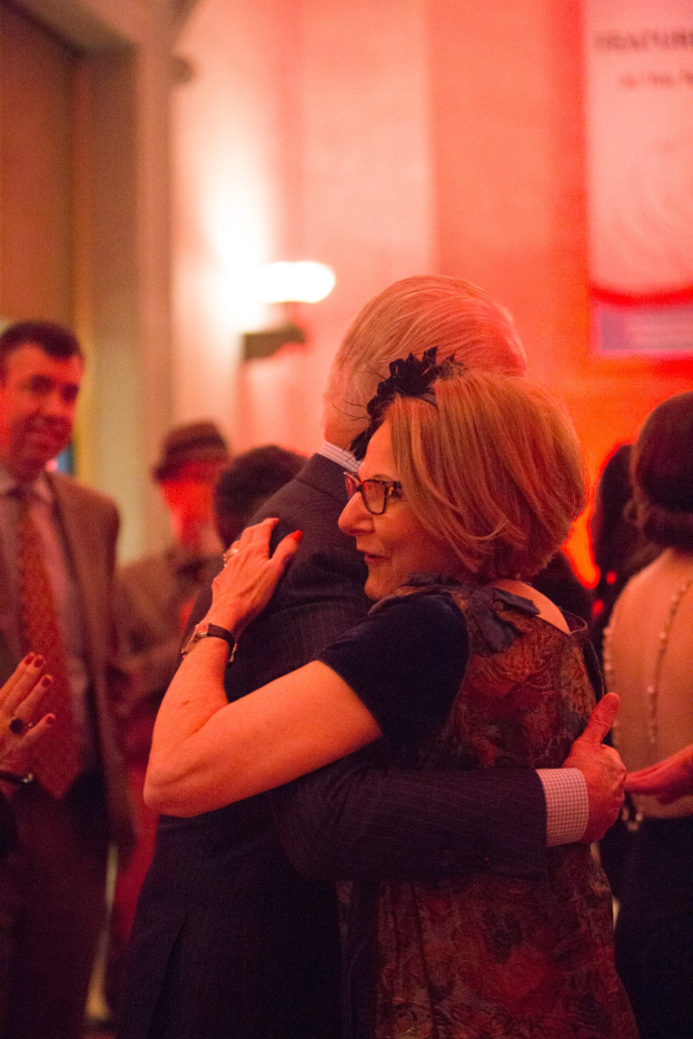 Zwei Menschen umarmen sich herzlich auf einer Party, umgeben von anderen im Hintergrund mit einer beleuchteten Wand dahinter.