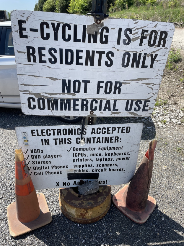 Schild mit der Aufschrift "E-Bike-Fahren nur für Anwohner" neben einer Straße mit Absperrgittern, einem Fahrzeug, Bäumen und einem klaren blauen Himmel im Hintergrund.