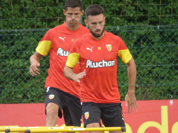 Zwei Männer in roten T-Shirts und schwarzen Shorts, die auf einem Feld mit einem Zaun und Bäumen im Hintergrund Fussball spielen, und ein rotes Schild mit 'Borussia Dortmund' darauf.