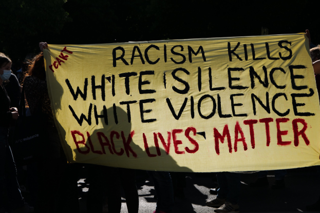 Gruppe maskierter Menschen mit einem "Rassismus tötet Weiße Stille, Weiße Gewalt, Black Lives Matter"-Schild auf einer dunklen Straße.